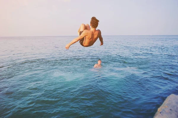 Young man jumping off cliff into blue water ocean at sunset. Active outdoor, holiday adventure, tourism action, healthy summer joy, Fun activity lifestyle. Crazy adult guy in swimwear fly from climb