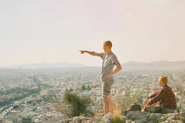 Pareja hombre y mujer en la cima de la montaña disfrutando de hermoso paisaje urbano Atenas Grecia en el fondo. Los turistas se relajan en la colina Atenas en verano. Famosa ciudad de Atenas en Europa. aventura de viaje . —  Fotos de Stock