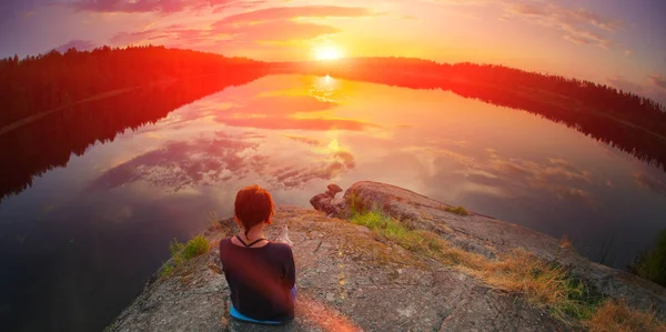 Young woman sitting enjoying peaceful moment of beautiful colorful sunset. In reflection of the lake water sees clouds and sun. Concepts of wellness happiness, peace freedom, inner mind meditating.