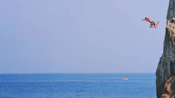 Młody człowiek skoki od urwisko w błękitne wody oceanu o zachodzie słońca. Ustawy — Zdjęcie stockowe