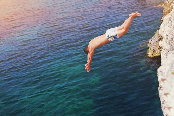 Young man jumping off cliff into blue water ocean at sunset. Act