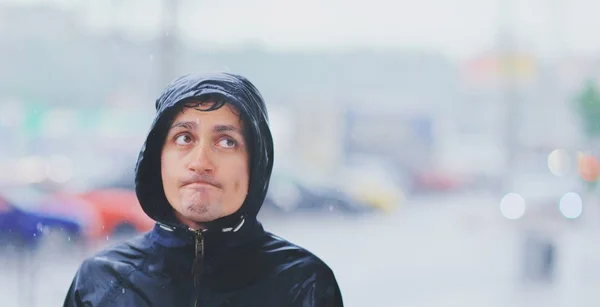 Retrato de um jovem molhado em uma jaqueta com um capuz na chuva no fundo borrado rua da cidade em tsunami, close-up. Mau tempo conceito pessimismo, frio outono infeliz ironia pessoas na capa de chuva . — Fotografia de Stock