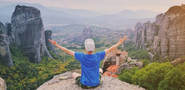 Man rise hand up sitting on top of mountain enjoying peaceful mountain landscape. Belief, praise, religion and worship. Freedom meditation and travel concept. Monastery Meteora Kalambaka in Greece.