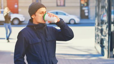 Yakışıklı genç adam gözlük takıyor ve elinde tek kullanımlık bardakla geziyor. Autumn City caddesinde kahve içiyor, şehir hayatının tadını çıkarıyor ve kışın açık havada sıcak kahvenin tadını çıkarıyor.