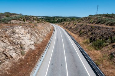 Dolambaçlı bir yol, kayalık yamaçlar ve berrak bir gökyüzünün altında seyrek bir bitki örtüsüyle çevrili kırsal bir arazide esiyor.