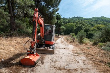 Mini kazıcı orman tarafından kuşatılmış kırsal bir yolun kenarlarını temizlemeye çalışıyor.