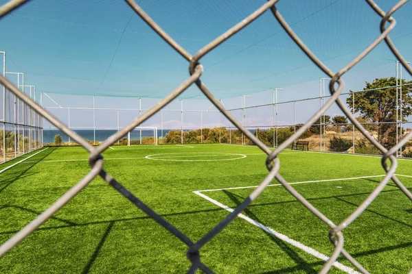 View of an outdoor green soccer pitch from behind a metal fence, near the sea.