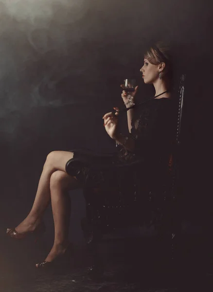Una chica en un vestido negro está sentada en una silla con una copa de vino — Foto de Stock