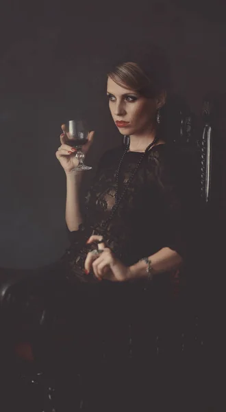 Una chica en un vestido negro está sentada en una silla con una copa de vino —  Fotos de Stock