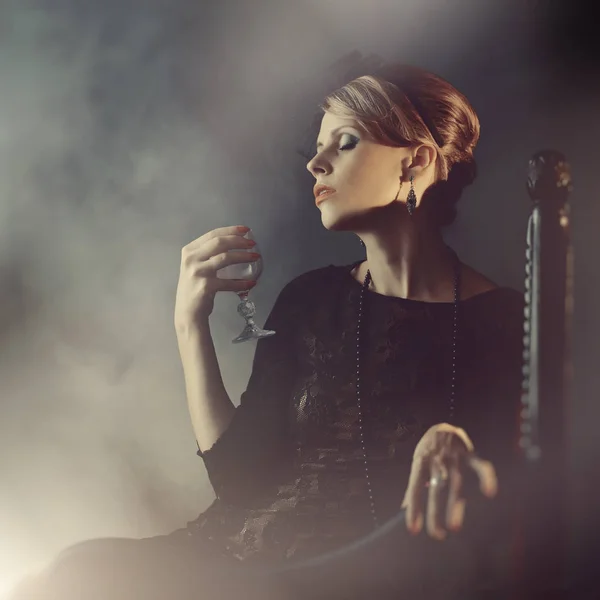 Una chica en un vestido negro está sentada en una silla con una copa de vino — Foto de Stock