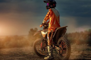 Rear view of a professional motorcyclist preparing before riding in the mountains and then on the off-road. This is a sunset. clipart