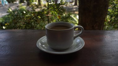 Dumanı tüten kahveyle dolu beyaz bir seramik fincan, tabağının üzerinde, koyu ahşap bir yüzeye oturur. Arka planda, yemyeşil yaprakları ve ağaçları olan yemyeşil bir bahçenin yumuşakça bulanık bir görüntüsü var.,