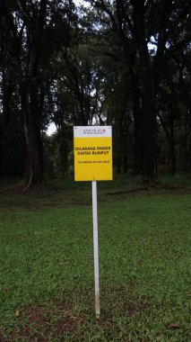 Ormanın yoğun yeşillikleriyle çevrili canlı yeşil bir çimenliğin ortasındaki beyaz bir direğin üzerinde sarı ve beyaz bir tabela duruyor. İşaret, hem Endonezce hem de İngilizce, açıkça ifade ediyor ki