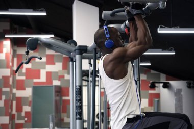 A black muscular guy in a white T-shirt and headphones pulls himself up on the simulator. Horizontal frame on the background of the gym clipart