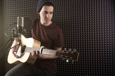 Koyu T-shirt giyen mikrofon önünde oturur ve güneşte gitar çalar. Yatay fotoğraf