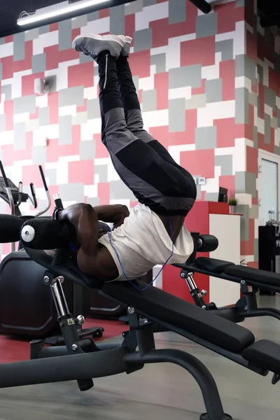 A black, muscular guy in a white T-shirt and headphones picks up his legs while lying on the simulator. Vertical side photo