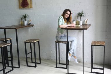 Kız yüksek masada kahve ile beyaz tuğla duvara kafasını kamera doğru dönerek oturur. Yatay fotoğraf