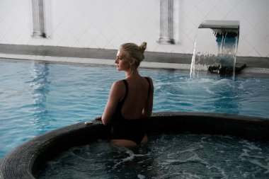 Young blonde woman in a swimsuit with open back stands waist-deep in the pool, turning her head in profile Horizontal photo clipart