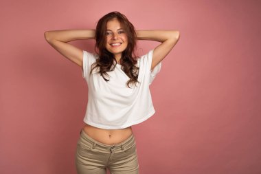 Young brunette in a white t-shirt and beige pants smiles broadly at the camera touching her head with both hands, pink background clipart