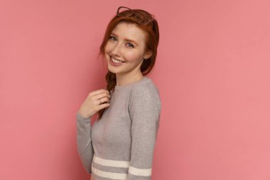 Redhead kız kamera biraz yan duruyor, ona örgü dokunur ve pembe arka planda kamera gülümsüyor