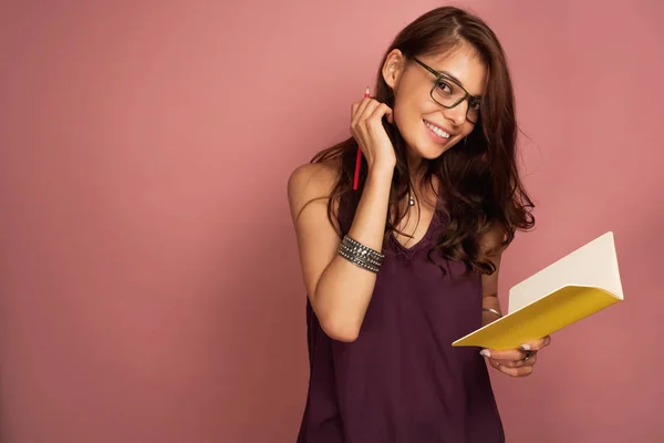 Ung mörkhårig kvinna tittar på kameran genom hennes glasögon med leende håller hennes gula antecknings bok, rosa bakgrund — Stockfoto