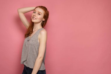 Redhead kız kameraya hafifçe yana duruyor, başının arkasına dokunur ve kameraya gülümsüyor