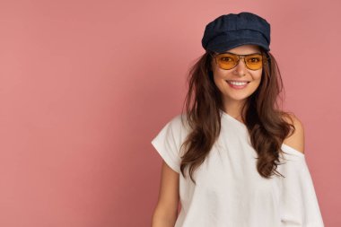 Sarı gözlüklü ve koyu şapkalı genç koyu saçlı kadın kameraya gülümsüyor, pembe arka plan