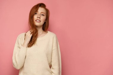 Genç kızıl saçlı kadın saçlarına dokunur ve pembe arka plan üzerinde kameraya bakar