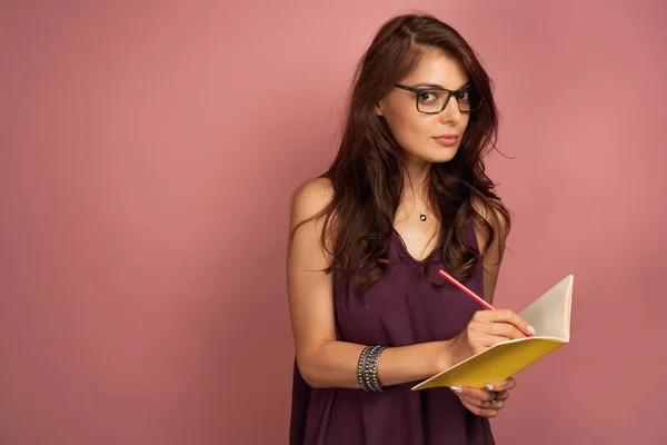 Young dark-haired woman writes something down in her notebook looking at the camera through her eyeglasses, pink background