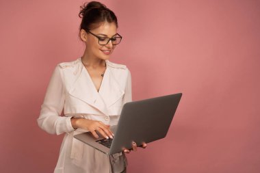 Beyaz önlük lü ve gözlüklü iş kadını dizüstü bilgisayarıyla gülümseyen, pembe arka planlı çalışıyor