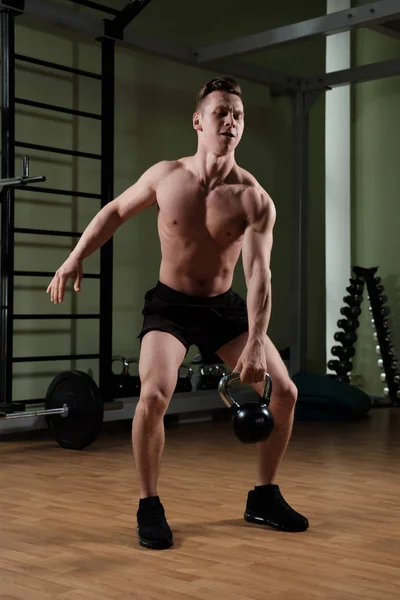 Un tipo musculoso con un torso desnudo en pantalones cortos deportivos se para en el gimnasio y levanta un kettlebell . — Foto de Stock