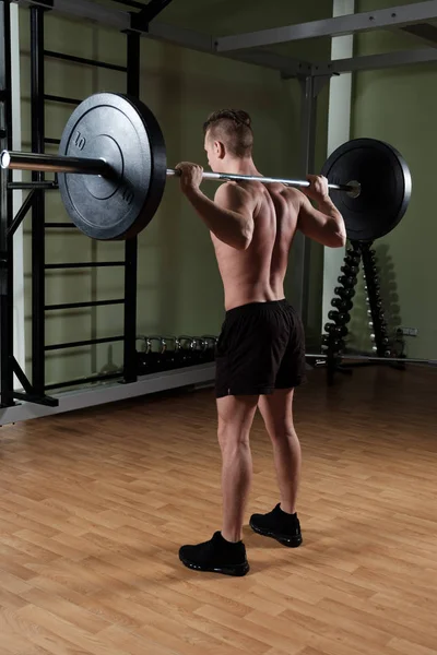 Ragazzo muscoloso con un busto nudo in pantaloncini sportivi con un bilanciere sulle spalle, una cornice dalla parte posteriore . — Foto Stock