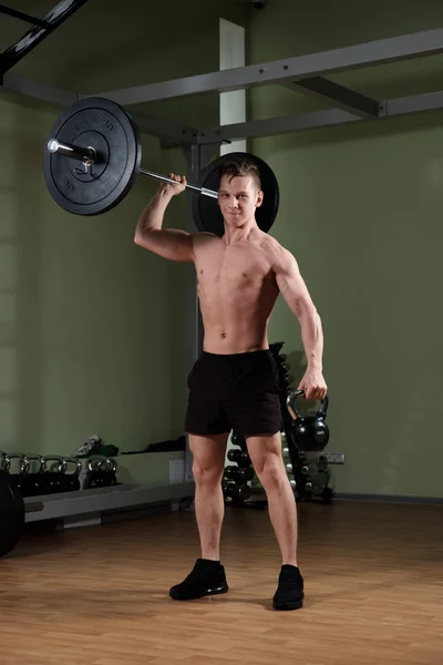 Un tipo musculoso con un torso desnudo en pantalones cortos deportivos levanta la barra con una mano y mira a la cámara . —  Fotos de Stock