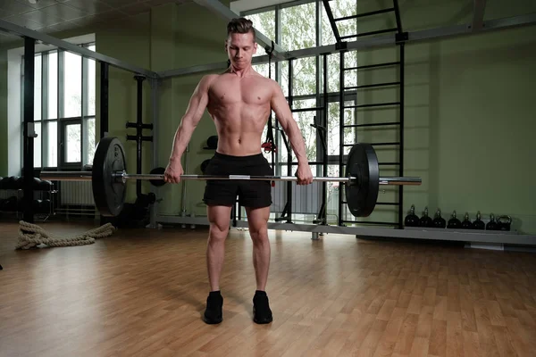 Un tipo musculoso en pantalones cortos deportivos y un torso desnudo está de pie en el gimnasio y está sosteniendo firmemente una pesada barra . —  Fotos de Stock
