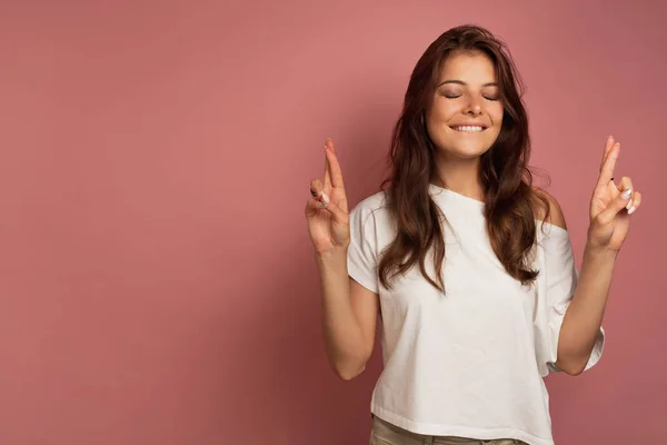 Ein Mädchen in weißem Oberteil steht auf rosa Hintergrund, schielt und drückt die Daumen. — Stockfoto