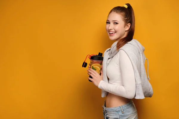 Roodharige meisje staande op een gele achtergrond in profiel met een sport shaker en glimlachend op de camera. — Stockfoto