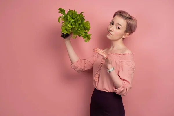 En flicka med kort hår i en blus, håller en sallad, pekar med handen och är skeptisk till kameran, på en rosa bakgrund — Stockfoto