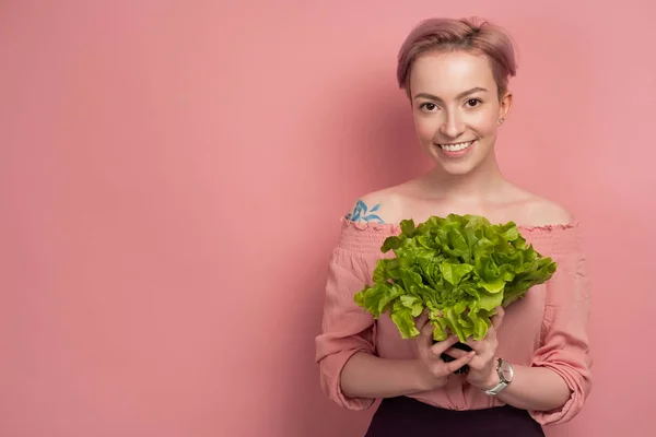 En tjej med kort rosa hår leende tittar på kameran, håller en sallad nära hennes bröst, på en rosa bakgrund. — Stockfoto