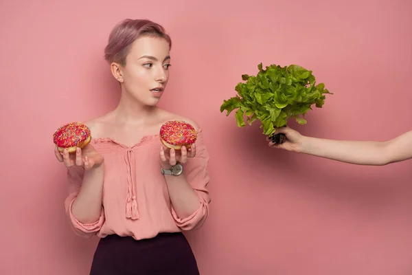 En tjej med kort hår i en blus håller munkar i händerna, och tittar på sallad i utsträckt hand, på en rosa bakgrund. — Stockfoto