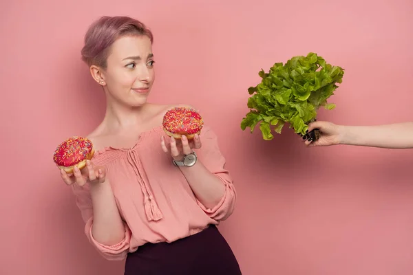 En tjej med kort rosa hår, håller munkar i händerna, fruktansvärt ta av från hennes händer med en sallad, på en rosa bakgrund. — Stockfoto