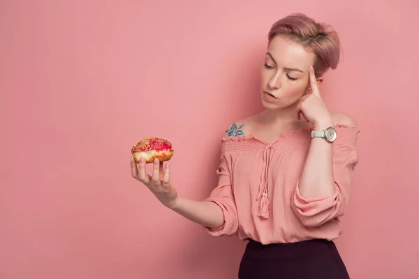 블라우스에 짧은 분홍색 머리를 가진 소녀는 분홍색 배경에, 머리에 그녀의 손가락을 넣어, 손에 도넛에 신중하게 보인다. — 스톡 사진