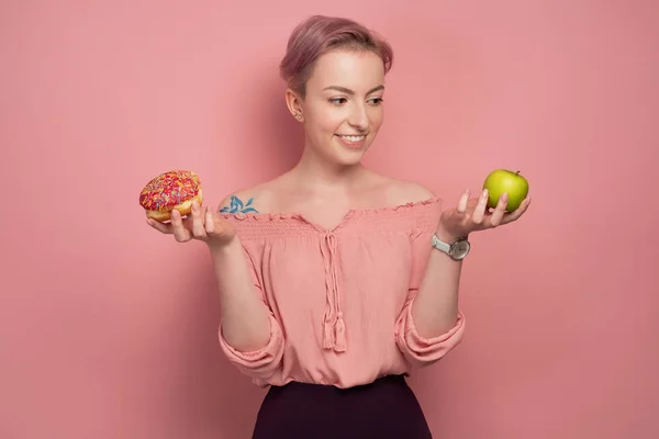 En tjej med kort rosa hår, håller en munk och ett grönt äpple i händerna, och tittar på äpplet med ett leende, på en rosa bakgrund. — Stockfoto