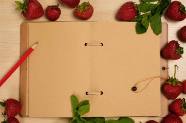 Kırmızı kalemile kraft kağıt açık bir defter hafif ahşap bir masa üzerinde çilek ve nane ile çevrilidir.