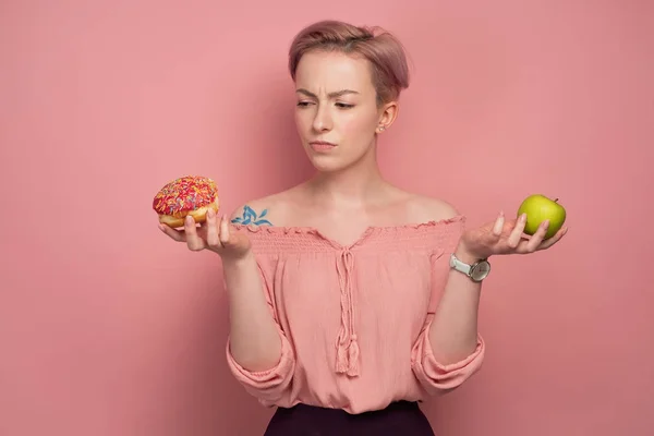 En tjej med kort rosa hår i en blus, håller ett grönt äpple och en munk i händerna, ser tankfullt på det, på en rosa bakgrund — Stockfoto