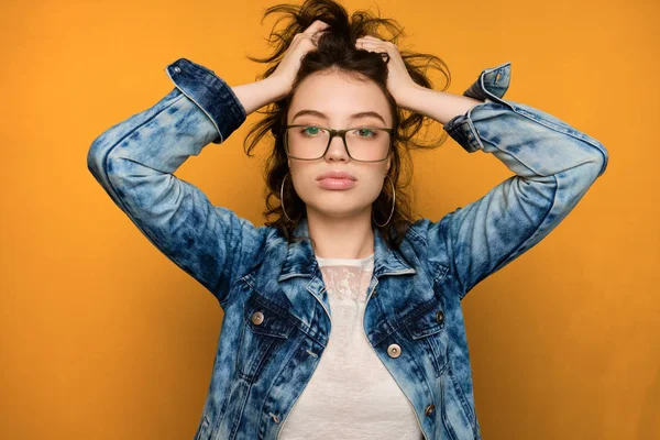 Uma menina encaracolada de cabelos escuros em jeans e óculos fica em um fundo amarelo, olhando para a câmera com a língua para fora e as mãos nos cabelos. . — Fotografia de Stock