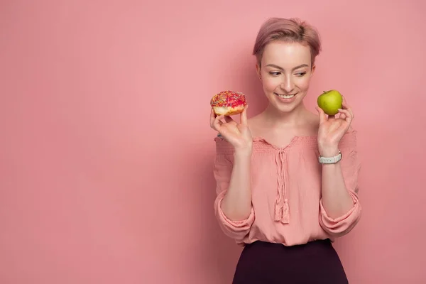 En tjej med kort rosa hår, håller en munk och ett grönt äpple nära hennes ansikte, och tittar på äpplet, leende, på en rosa bakgrund. — Stockfoto