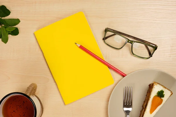 Yellow notepad with a red pencil and glasses, with a mug of tea and a piece of cake lie on a wooden table