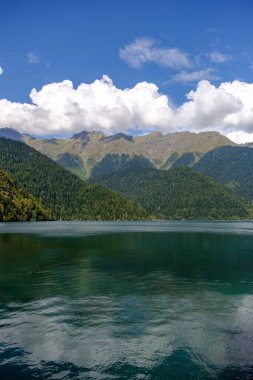 Abhazya'da Ritsa gölü ve dağları. Dağların üzerinde mavi gökyüzünde beyaz bulutlar.