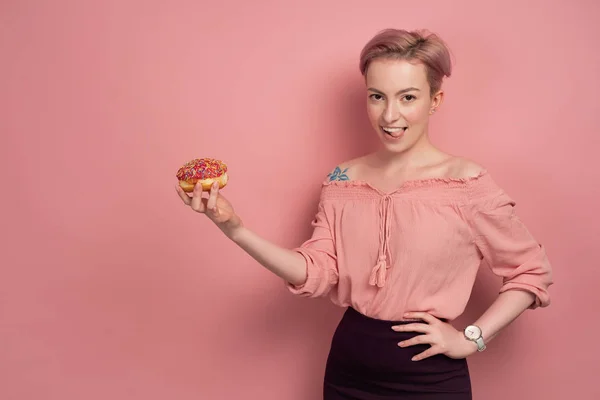 짧은 분홍색 머리를 가진 소녀, 그녀의 손에 도넛을 잡고, 그녀의 혀를 물고 카메라를 보고, 분홍색 배경에. — 스톡 사진
