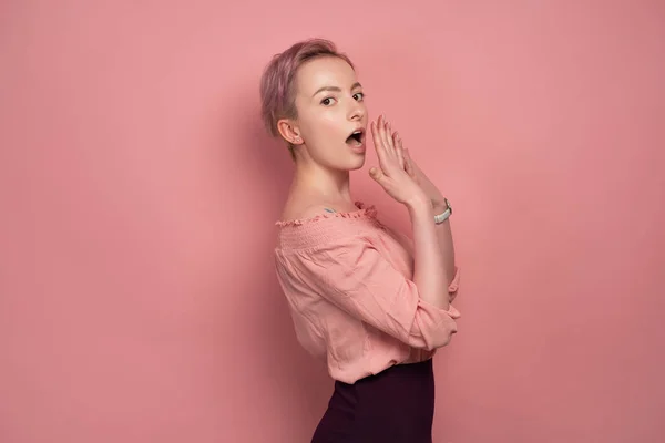 Egy lány, rövid haj áll profilja a rózsaszín háttér és nézte a kamerát egy zavarba a száj fedezetű — Stock Fotó